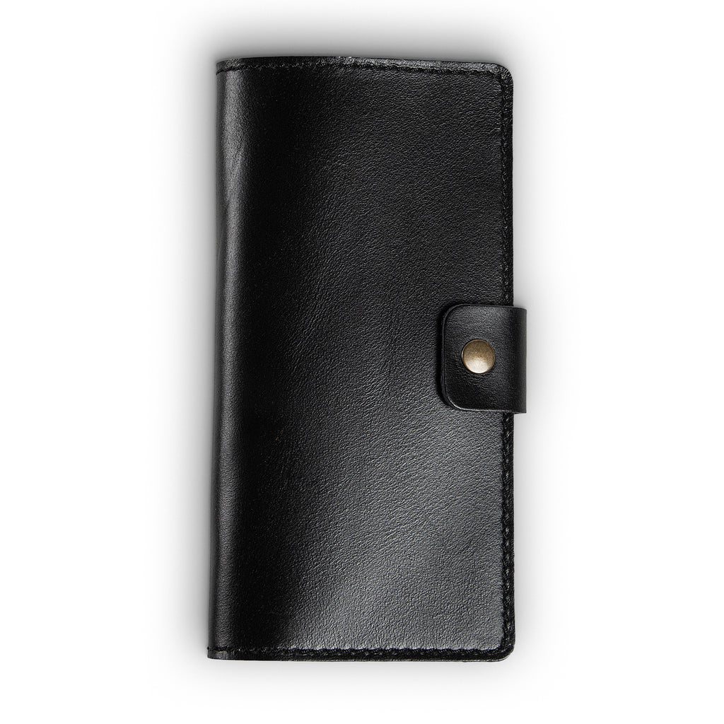Clark Leather Checkbook Cover in Dexios Black, closed and laid flat on a white surface. Premium top-grain Dexios calfskin in a deep glossy black with smooth pull-up surface sheen and subtle natural grain variation. No visible exterior stitching on this colorway. A small leather tab with an antique brass snap closure sits centered on the right edge.