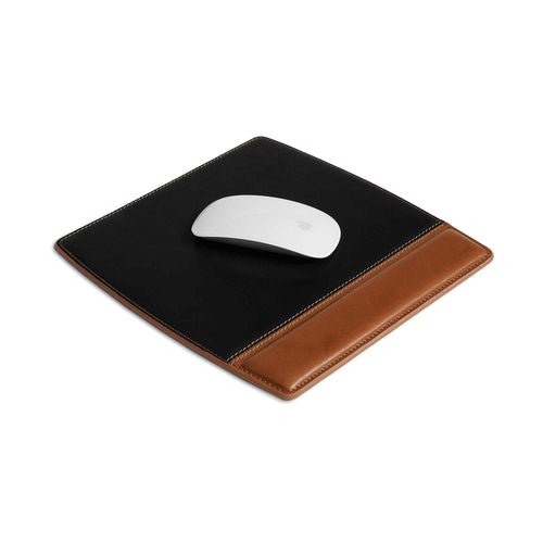 Bumpy Leather Mouse Pad in Maui Charcoal Black laid flat at an angle on a white surface with a white Apple Magic Mouse resting on the suede upper panel. The two-tone construction is clearly visible — matte black suede tracking surface above, warm cognac tan leather wrist rest below — with contrast saddle stitching along all edges.