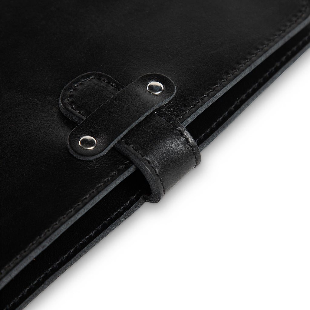 Close-up of DuPage Black journal's leather strap closure on Dexios calfskin, showing two silver-tone rivets, saddle-stitched strap loop, and black edge stitching detail.