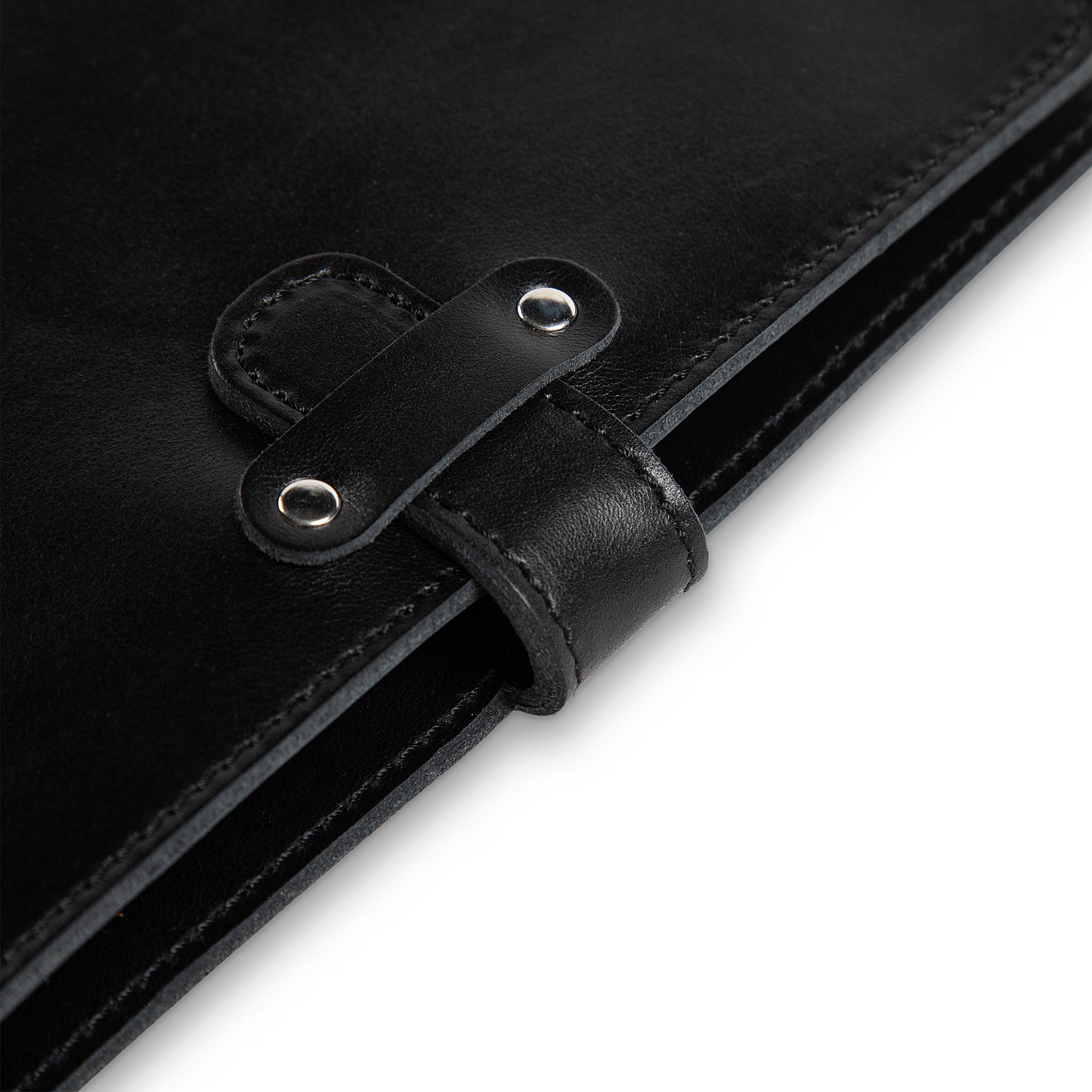 Close-up of DuPage Black journal's leather strap closure on Dexios calfskin, showing two silver-tone rivets, saddle-stitched strap loop, and black edge stitching detail.