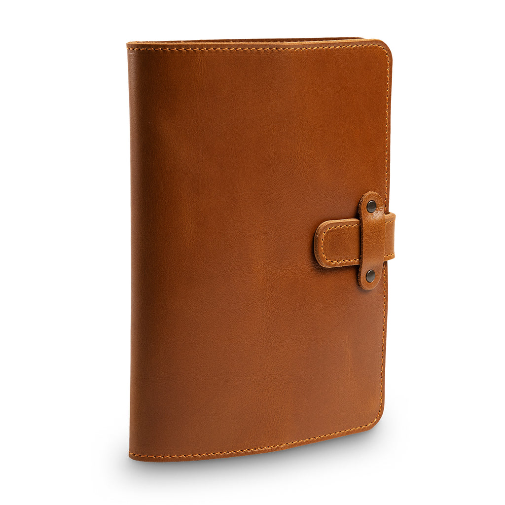 Three-quarter front view of closed mustard brown DuPage journal showing spine thickness, orange saddle stitching, side strap closure tab, and embossed PEGAI logo on lower front cover.