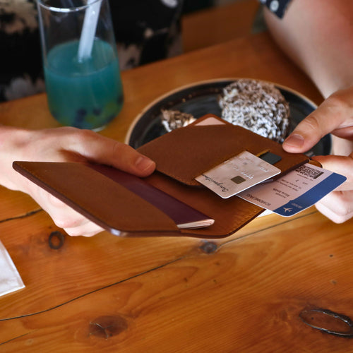 Hands holding an open cinnamon tan leather passport wallet at a wood café table. One hand holds the wallet open while another pulls a boarding pass from the card pocket. A credit card is also visible. Warm pebbled exterior and amber tan interior lining. Drink and dessert in background.