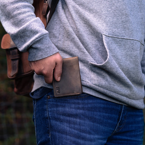A person wearing a gray sweatshirt and dark indigo jeans pulling the Bureau Leather Bifold Wallet in Chestnut Brown from their back pocket. Fire-branded initials "JPV" visible on the lower front face. The wallet's warm chestnut Dallas Crazy Horse calfskin shows rich pull-up tonal variation. A tan leather bag strap visible over the shoulder, with blurred outdoor greenery in the background.