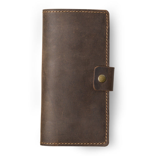 Clark Leather Checkbook Cover in Chestnut Brown, closed and laid flat on a white surface. Dallas Crazy Horse calfskin in a warm deep chestnut tone with natural waxy pull-up surface variation and visible hide markings. Warm tonal saddle stitching runs along all four edges. A small leather tab with an antique brass snap closure sits centered on the right edge.