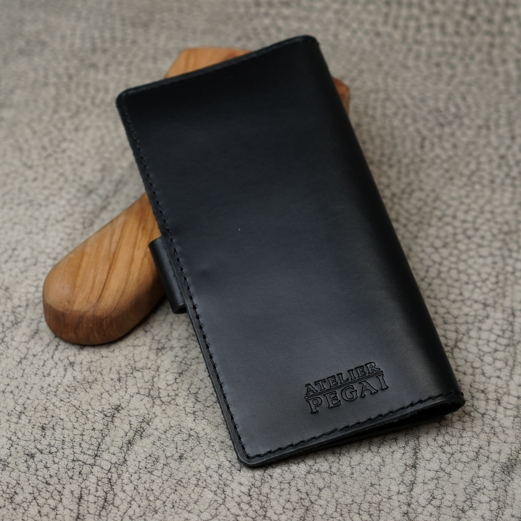 Clark Leather Checkbook Cover in Dual Tone Black propped upright against a smooth wooden tool handle on a cream crackled leather surface. Deep black DT Lux cowhide with a rich glossy pull-up finish reveals subtle dual-tone depth where the leather curves and bends. Debossed "Atelier PEGAI" wordmark centered on the lower portion of the front face. Tonal dark saddle stitching along all edges. Black leather snap tab visible on the left side.