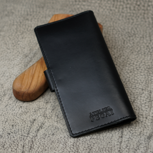 Clark Leather Checkbook Cover in Dual Tone Black propped upright against a smooth wooden tool handle on a cream crackled leather surface. Deep black DT Lux cowhide with a rich glossy pull-up finish reveals subtle dual-tone depth where the leather curves and bends. Debossed "Atelier PEGAI" wordmark centered on the lower portion of the front face. Tonal dark saddle stitching along all edges. Black leather snap tab visible on the left side.