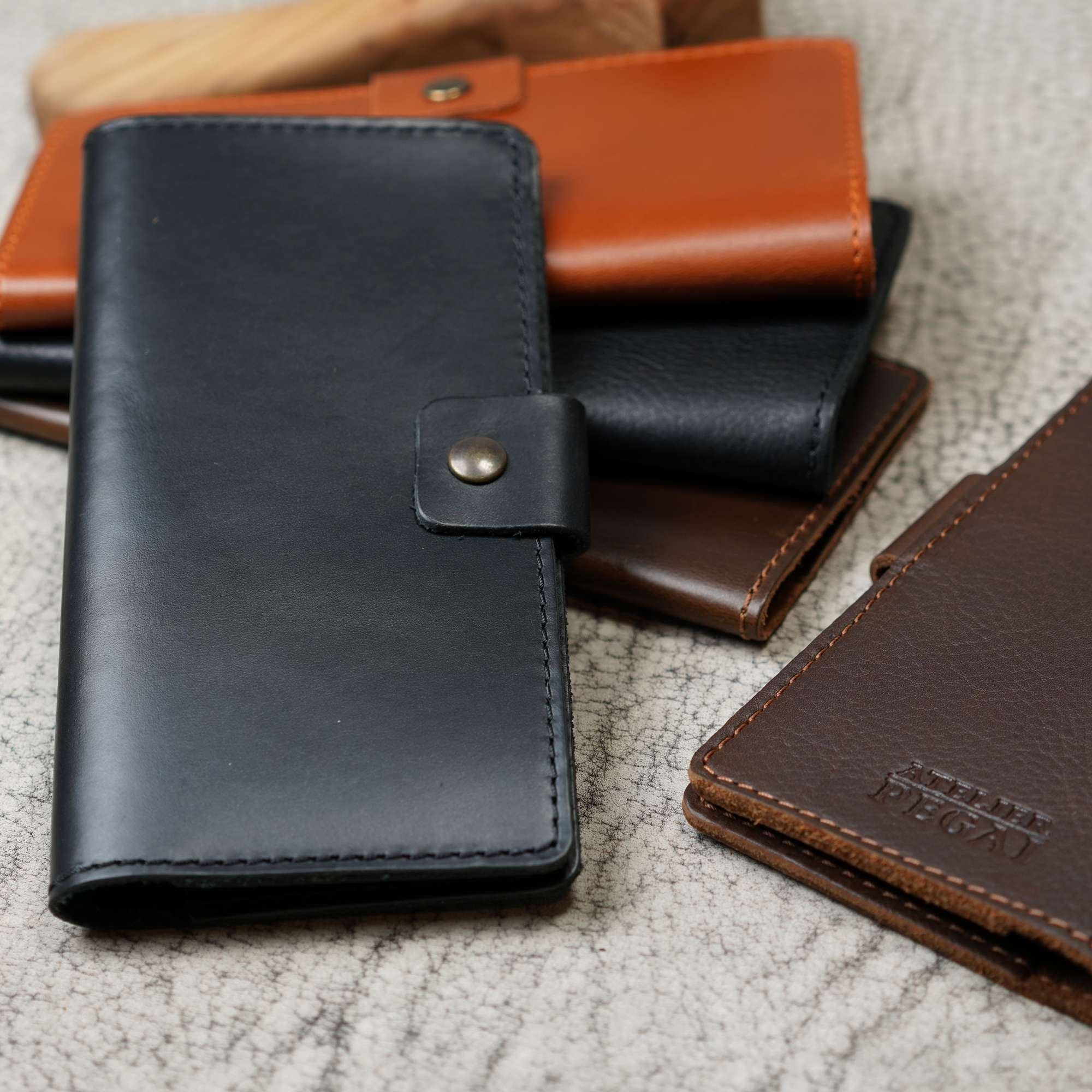 Clark Leather Checkbook Cover in Dual Tone Black styled in the foreground alongside multiple other Clark colorways on a cream crackled leather surface. The black cover with antique brass snap is most prominent. Behind and beside it, covers in tan cognac, dark brown, and additional black tones are layered together with a rustic wooden tool visible in the upper background. A dark brown cover in the lower right displays the debossed "Atelier PEGAI" logo. The grouping showcases the full range of Clark colorways