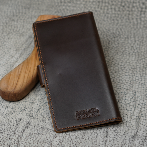 Clark Leather Checkbook Cover in Dual Tone Coffee propped upright against a smooth wooden tool handle on a cream crackled leather surface. Deep espresso DT Lux cowhide with a rich glossy pull-up finish shows warm dual-tone depth where the leather curves and catches directional light. Debossed "Atelier PEGAI" wordmark centered on the lower portion of the front face. Warm tonal brown saddle stitching along all edges. Dark leather snap tab visible on the left side.
