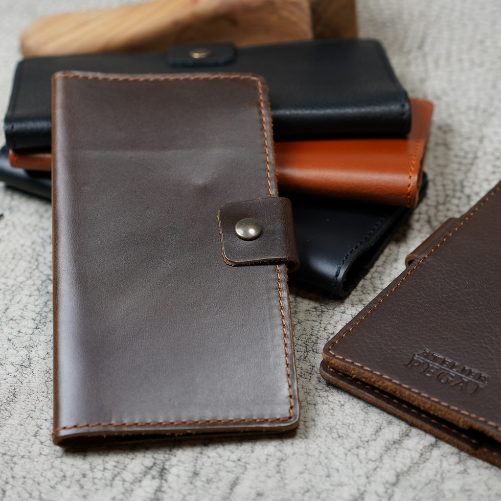 Clark Leather Checkbook Cover in Dual Tone Coffee shown with other Clark colorways — black, tan cognac, and dark brown — grouped on a crackled leather surface with a wooden tool. Dark brown cover at right shows debossed Atelier PEGAI logo.