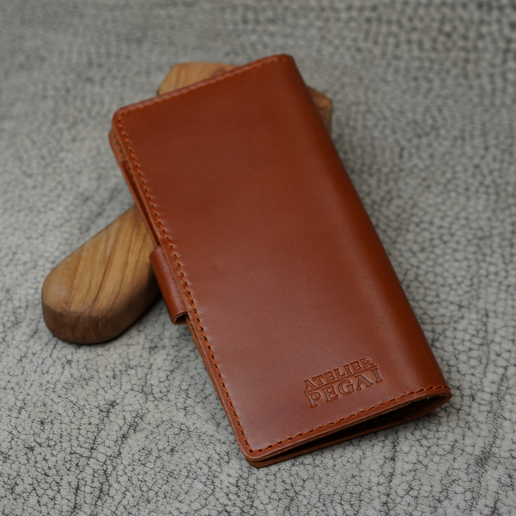 Clark Checkbook Cover in Dual Tone Whiskey propped on a wooden tool on crackled leather surface. Glossy warm tan DT Lux finish. Debossed "Atelier PEGAI" logo at lower front. Tonal stitching throughout.