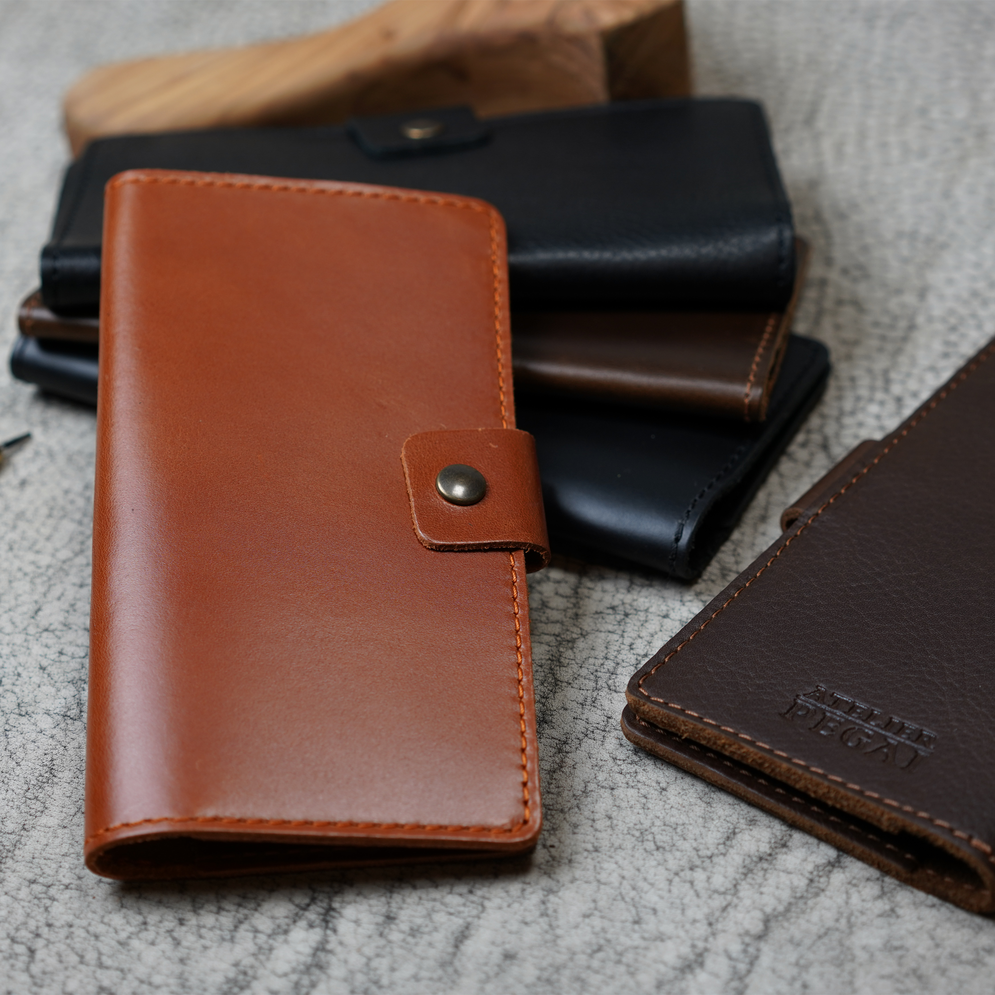 Clark Checkbook Cover in Dual Tone Whiskey in foreground with black, coffee, and dark brown colorways grouped on crackled leather. Dark brown cover at right shows debossed Atelier PEGAI logo.