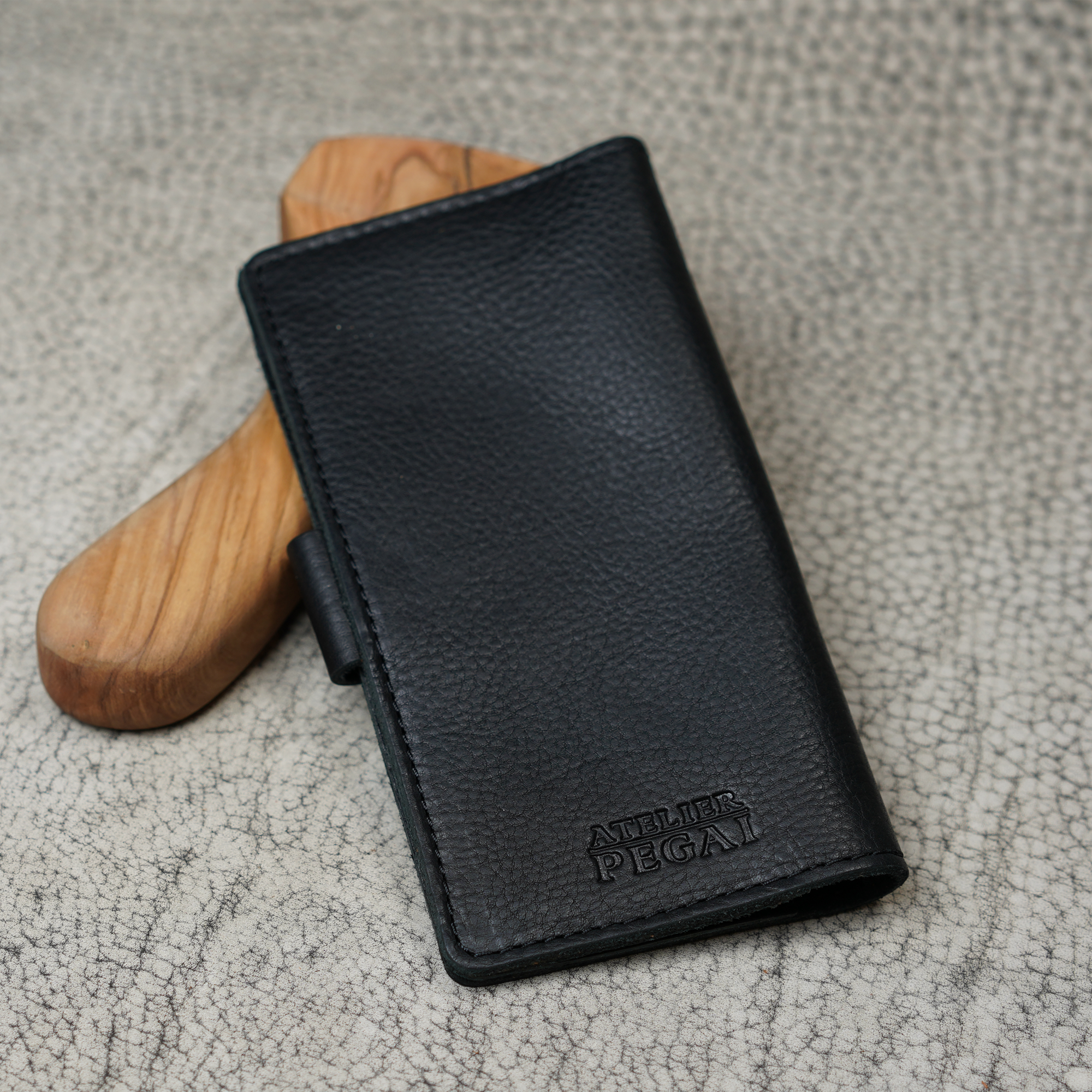 Clark Checkbook Cover in Dymnos Black propped on a wooden tool on crackled leather surface. Pebbled full-grain calfskin with rich pull-up depth. Debossed "Atelier PEGAI" logo at lower front.