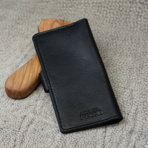 Clark Checkbook Cover in Dymnos Black propped on a wooden tool on crackled leather surface. Pebbled full-grain calfskin with rich pull-up depth. Debossed "Atelier PEGAI" logo at lower front.