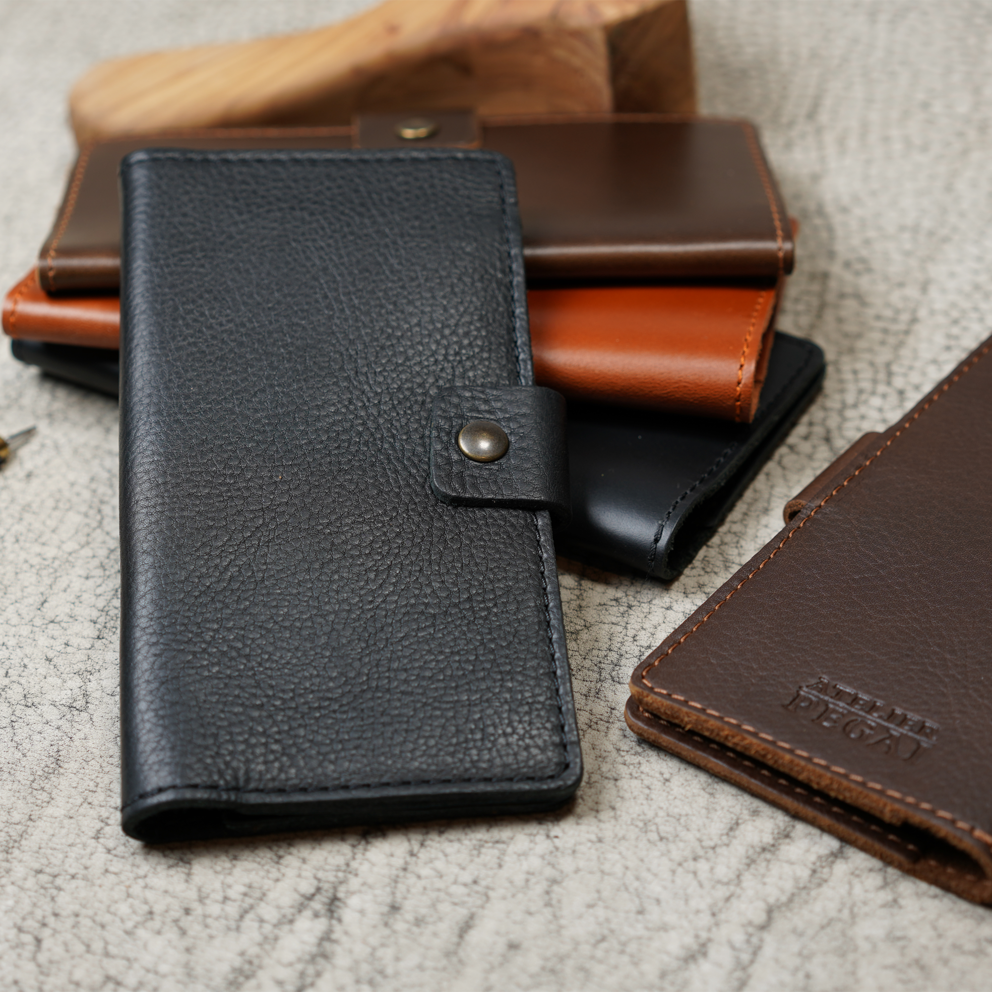 Clark Checkbook Cover in Dymnos Black in foreground with coffee, whiskey, black, and dark brown colorways grouped on crackled leather. Dark brown cover at right shows debossed Atelier PEGAI logo.