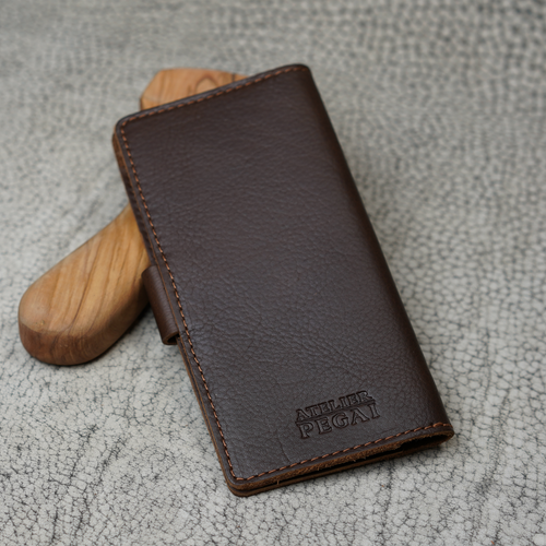 Clark Checkbook Cover in Dymnos Brown propped on a wooden tool on crackled leather surface. Deep brown pebbled full-grain calfskin. Debossed "Atelier PEGAI" logo at lower front. Warm tonal stitching.