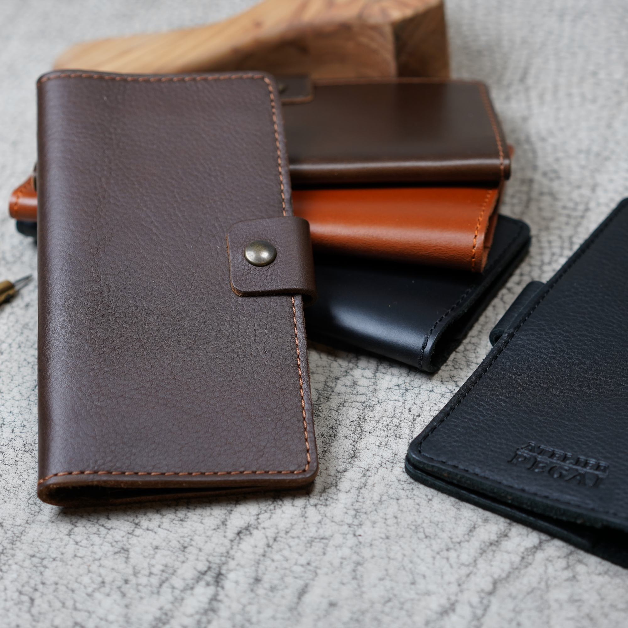 Clark Checkbook Cover in Dymnos Brown in foreground with whiskey, black, and dark brown colorways on crackled leather. Black cover at right shows debossed Atelier PEGAI logo. Pen visible at left.