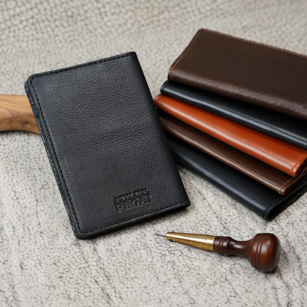 Black pebbled leather passport cover upright on a crackle-textured surface, Atelier PEGAI logo embossed at lower center. Behind it, a stack of four DeKalb covers in dark brown, black, cognac, and brown colorways. A wood-handled leather awl and driftwood piece rest alongside.