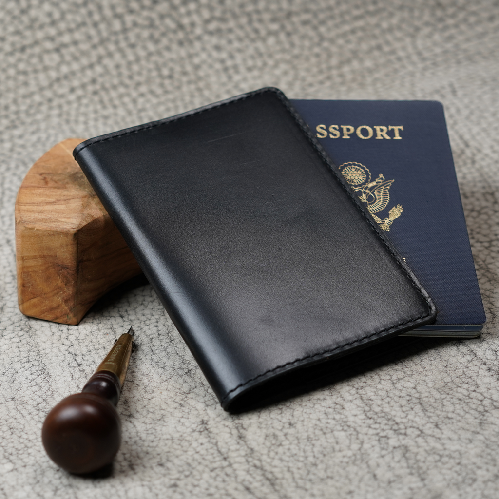 Black leather passport cover leaning against a small wood block beside a U.S. navy blue passport. A wood-handled leather awl rests in the foreground on a textured gray surface. Glossy dual-tone finish and black stitching visible on the cover's front panel.