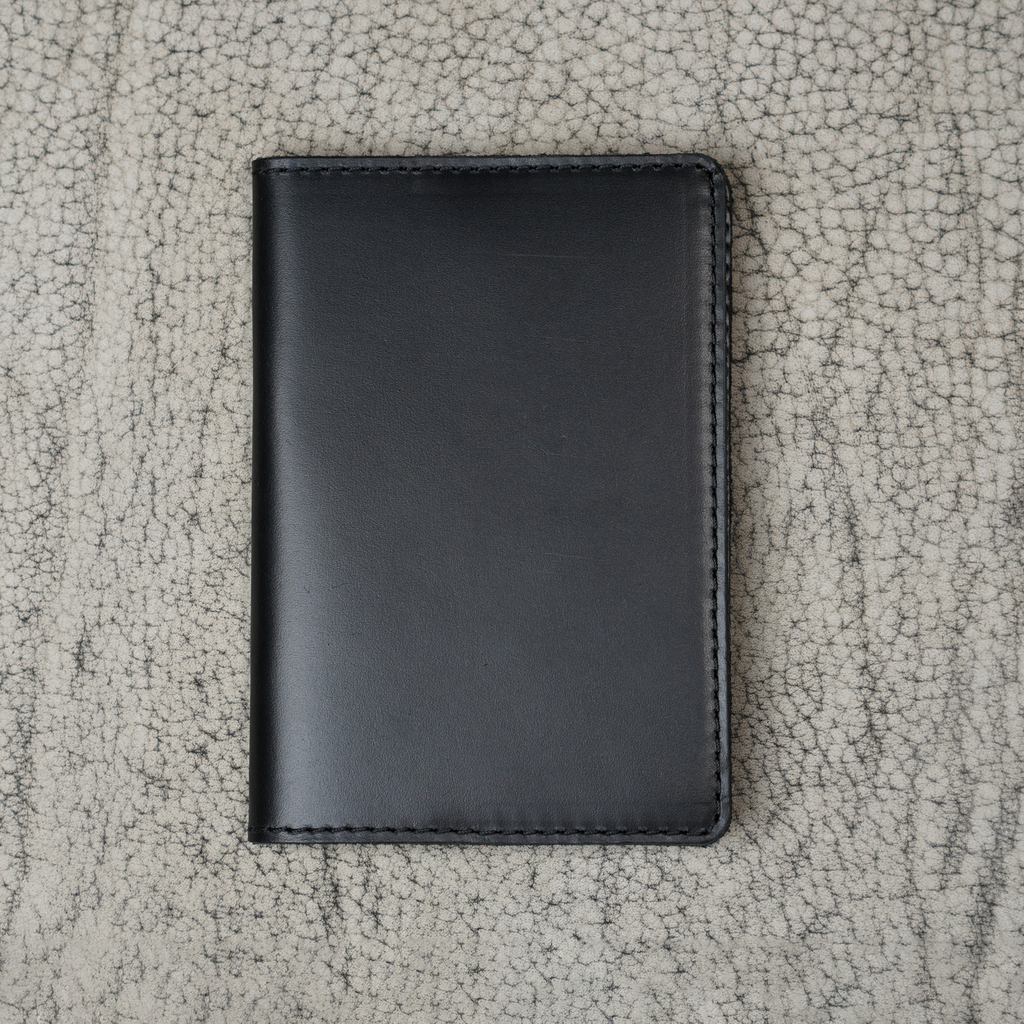 Black leather passport cover laid flat on a natural crackle-textured gray surface. Smooth glossy pull-up leather with subtle tonal depth. Hand-stitched black perimeter and rounded corners. Clean unadorned front panel with no hardware or closures.