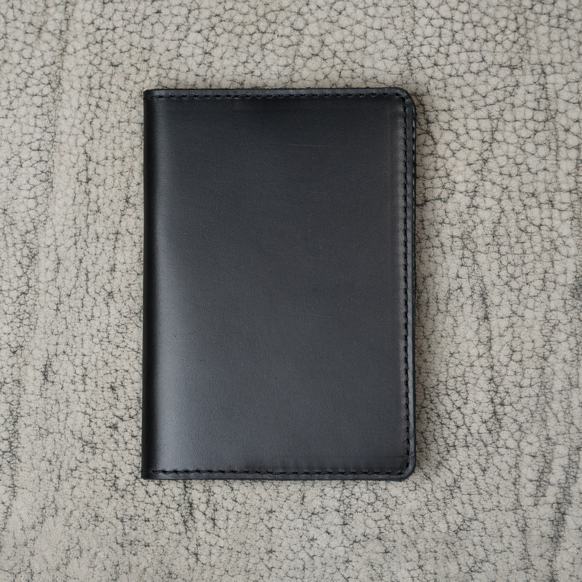 Black leather passport cover laid flat on a natural crackle-textured gray surface. Smooth glossy pull-up leather with subtle tonal depth. Hand-stitched black perimeter and rounded corners. Clean unadorned front panel with no hardware or closures.