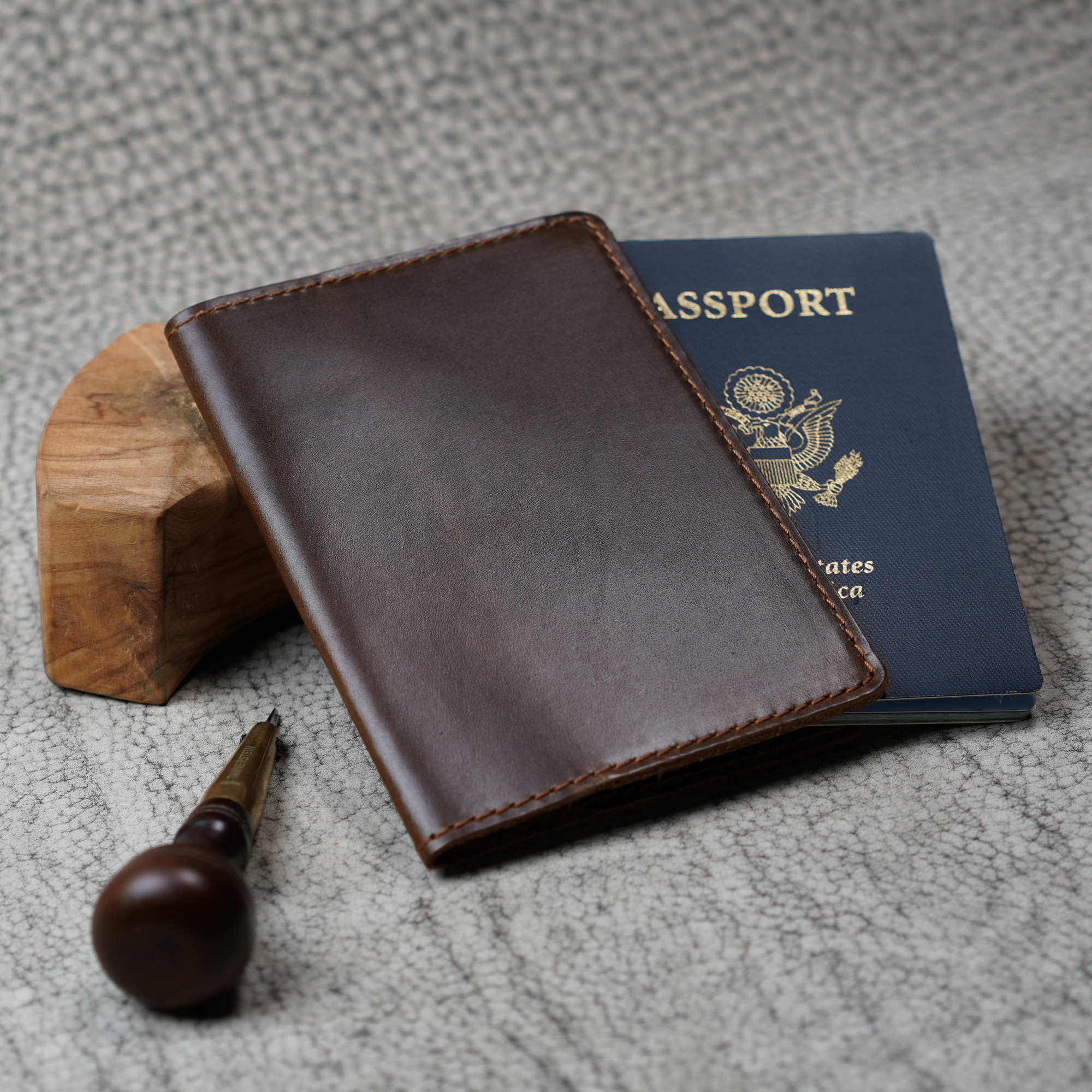 Deep brown leather passport cover leaning against a small wood block beside a U.S. navy blue passport. A wood-handled leather awl rests in the foreground on a textured gray surface. Glossy dual-tone coffee finish and brown saddle stitching visible on the cover's front panel.