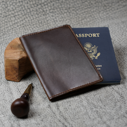 Deep brown leather passport cover leaning against a small wood block beside a U.S. navy blue passport. A wood-handled leather awl rests in the foreground on a textured gray surface. Glossy dual-tone coffee finish and brown saddle stitching visible on the cover's front panel.