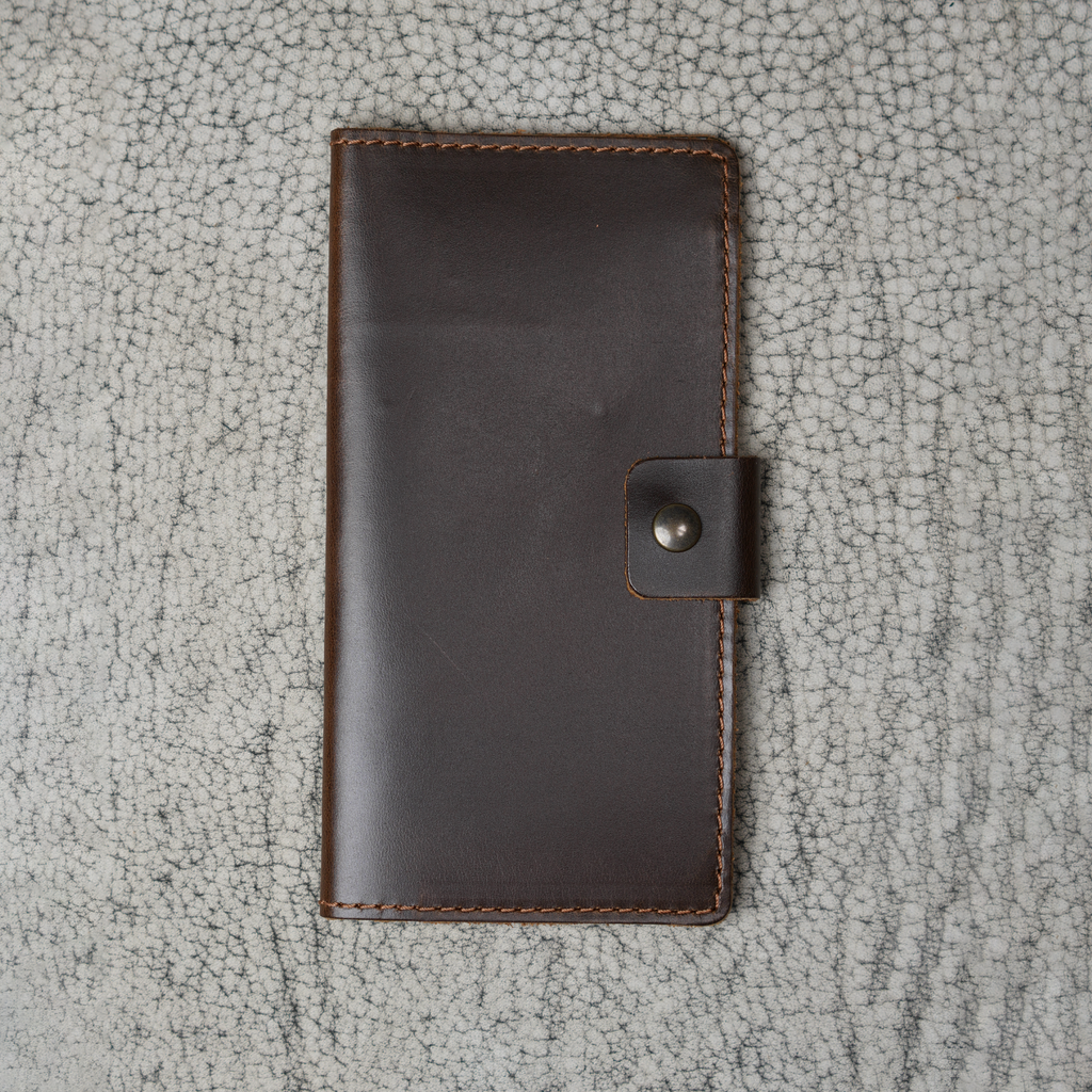 Clark Leather Checkbook Cover in Dual Tone Coffee laid flat on a cream crackled leather surface. Deep espresso DT Lux cowhide with a glossy pull-up finish shows rich tonal variation and natural light movement across the surface. Warm tonal brown saddle stitching runs along all four edges. Antique brass snap closure centered on the right edge. The editorial crackled surface highlights the depth and warmth of the coffee leather tone.