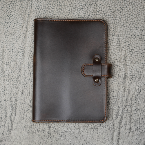 Dual Tone Coffee DT Lux leather journal closed on a cracked stone surface, showing glossy deep brown cowhide cover with dual-tone depth, tan stitching, and antique brass rivet strap closure.
