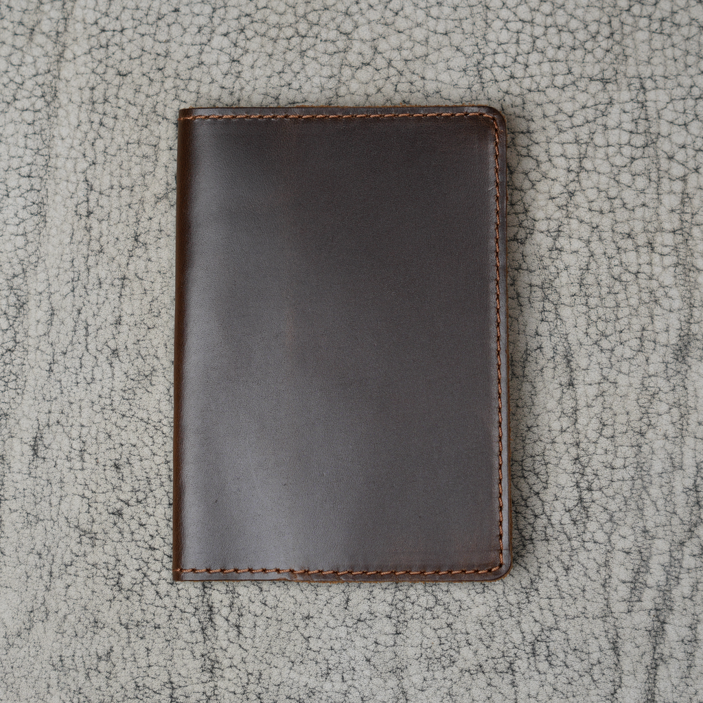 Dual tone coffee leather passport cover laid flat on a natural crackle-textured gray surface. Deep espresso brown glossy pull-up leather with warm tonal depth and subtle sheen. Hand-stitched brown perimeter and rounded corners. Clean unadorned front panel with no hardware.