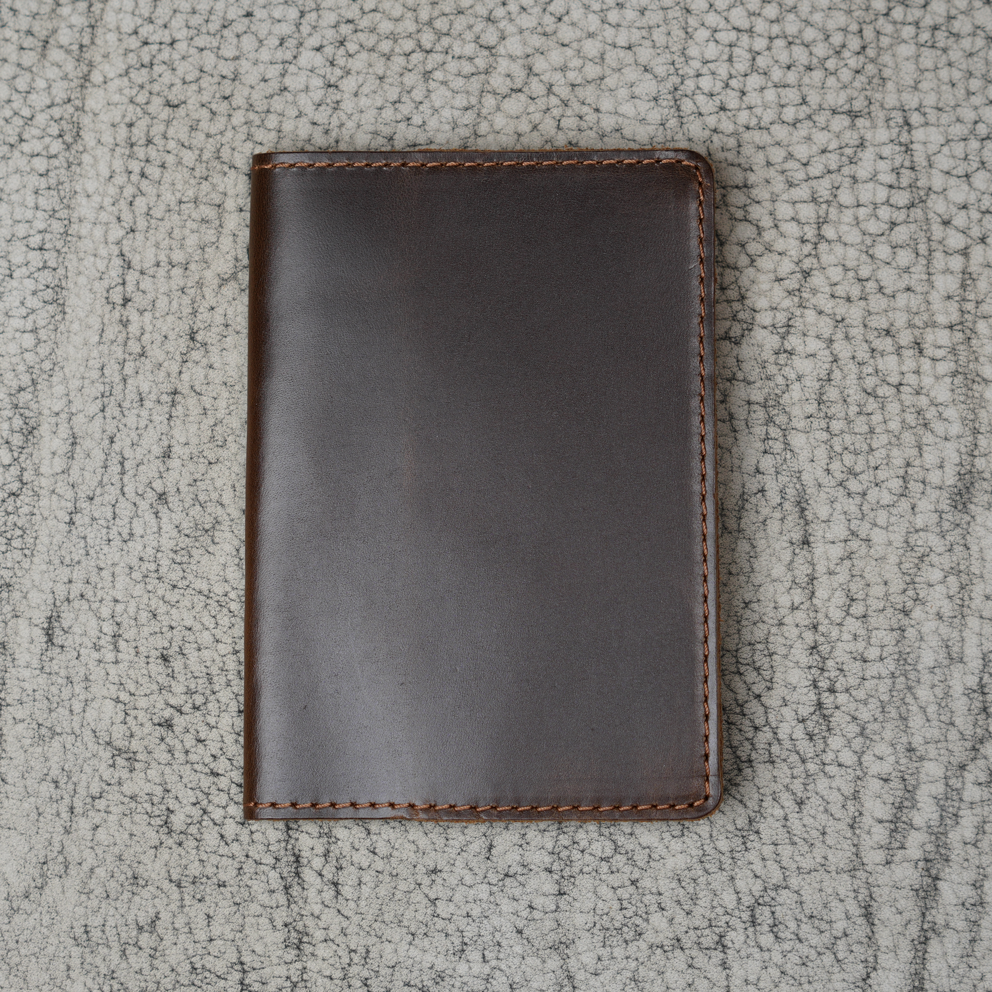 Dual tone coffee leather passport cover laid flat on a natural crackle-textured gray surface. Deep espresso brown glossy pull-up leather with warm tonal depth and subtle sheen. Hand-stitched brown perimeter and rounded corners. Clean unadorned front panel with no hardware.