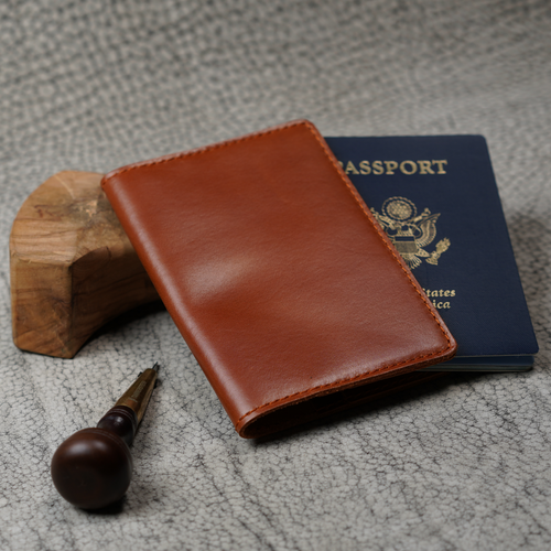 Warm cognac leather passport cover leaning against a small wood block beside a U.S. navy blue passport. A wood-handled leather awl rests in the foreground on a textured gray surface. Glossy dual-tone whiskey finish and orange saddle stitching visible on the front panel.