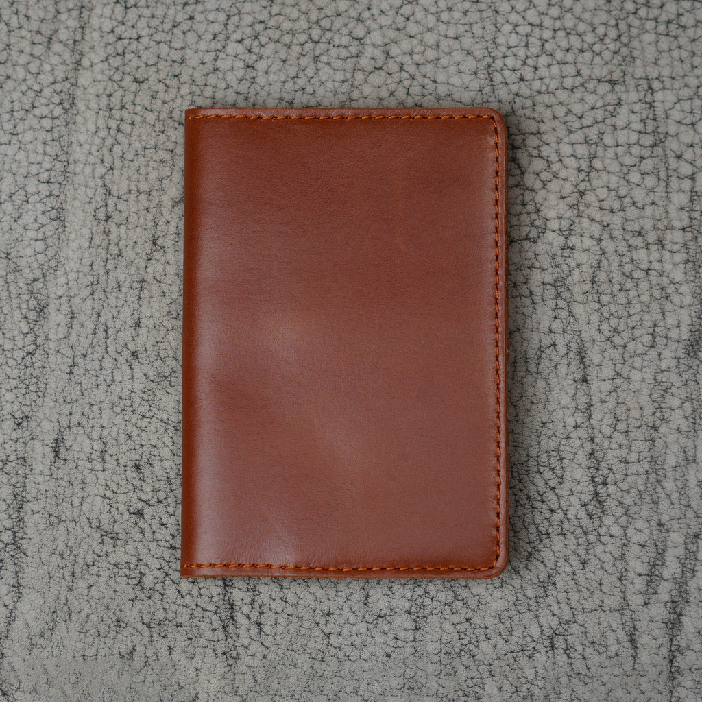 Dual tone whiskey leather passport cover laid flat on a natural crackle-textured gray surface. Warm cognac brown glossy pull-up leather with bright amber tonal movement and natural sheen. Hand-stitched burnt orange perimeter and rounded corners. Clean unadorned front panel.