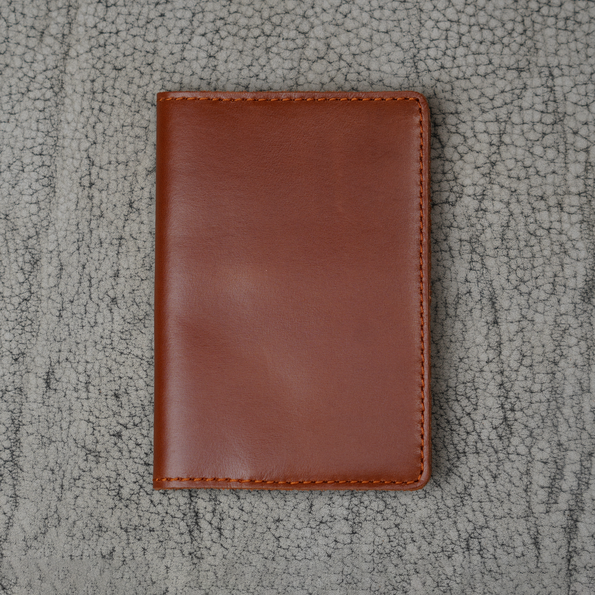 Dual tone whiskey leather passport cover laid flat on a natural crackle-textured gray surface. Warm cognac brown glossy pull-up leather with bright amber tonal movement and natural sheen. Hand-stitched burnt orange perimeter and rounded corners. Clean unadorned front panel.