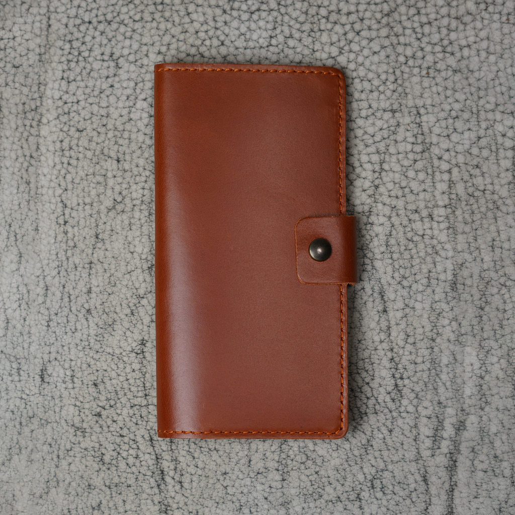 Clark Checkbook Cover in Dual Tone Whiskey laid flat on a cream crackled leather surface. Warm tan DT Lux cowhide with glossy pull-up sheen. Tonal saddle stitching on all edges. Antique brass snap on right.