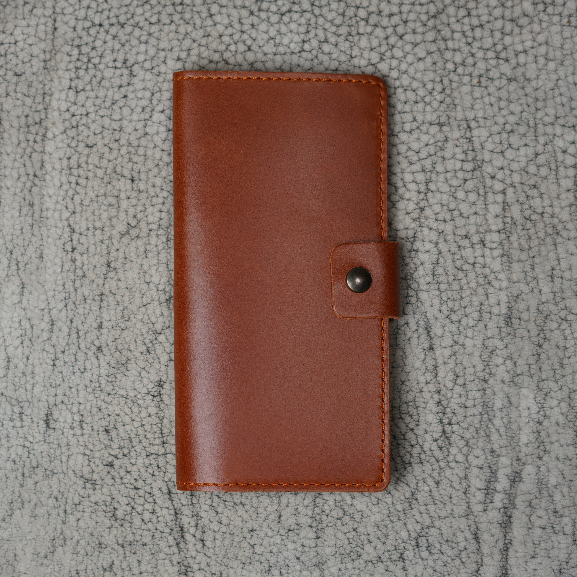 Clark Checkbook Cover in Dual Tone Whiskey laid flat on a cream crackled leather surface. Warm tan DT Lux cowhide with glossy pull-up sheen. Tonal saddle stitching on all edges. Antique brass snap on right.