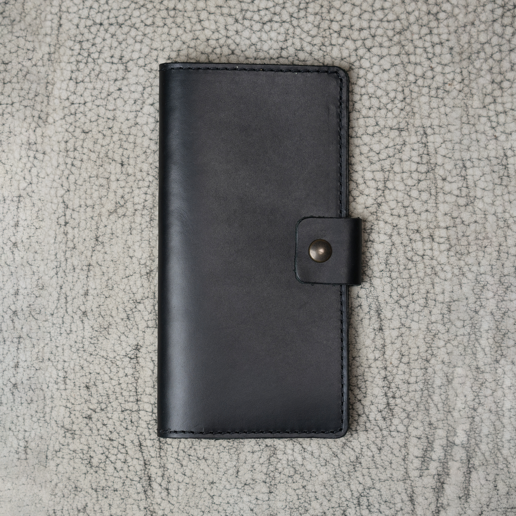 Clark Leather Checkbook Cover in Dual Tone Black laid flat on a cream crackled leather surface. Deep black DT Lux cowhide with a glossy pull-up finish shows subtle tonal depth and natural light variation across the surface. Tonal dark saddle stitching runs along all four edges. Antique brass snap closure centered on the right edge. The editorial surface reinforces the refined, understated luxury of the piece.