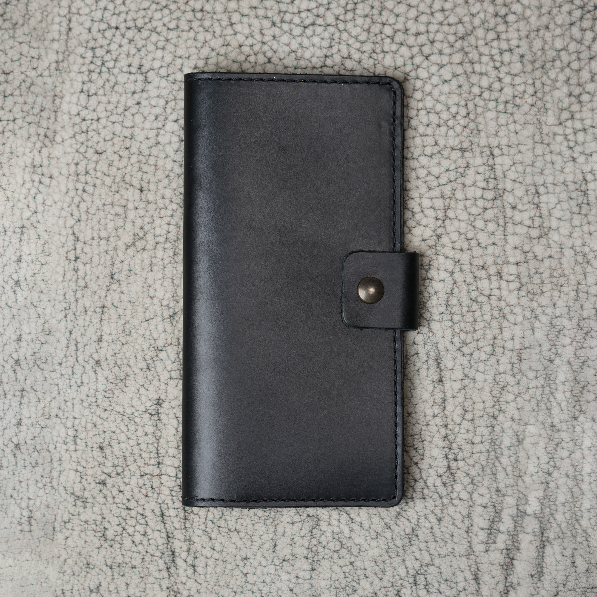 Clark Leather Checkbook Cover in Dual Tone Black laid flat on a cream crackled leather surface. Deep black DT Lux cowhide with a glossy pull-up finish shows subtle tonal depth and natural light variation across the surface. Tonal dark saddle stitching runs along all four edges. Antique brass snap closure centered on the right edge. The editorial surface reinforces the refined, understated luxury of the piece.