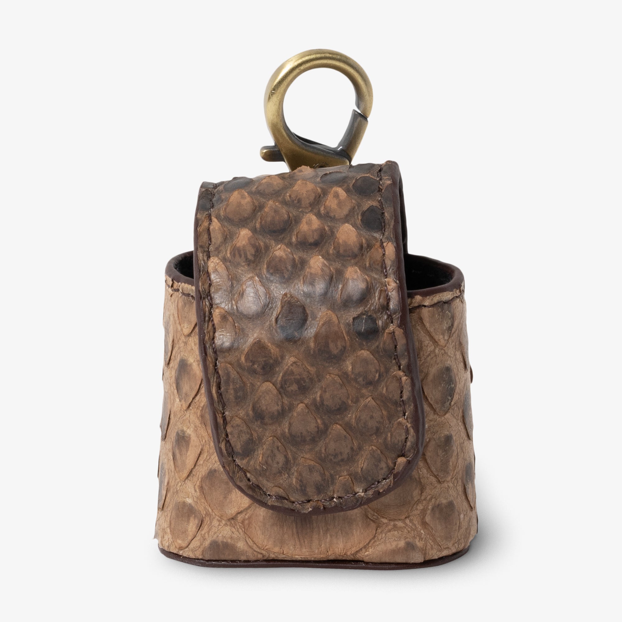 Coil AirPods case in Dune python leather, angled side view. Rounded scale texture in warm tan and brown tones. Curved magnetic flap with hand-stitched dark trim. Antique brass clip hardware at top.