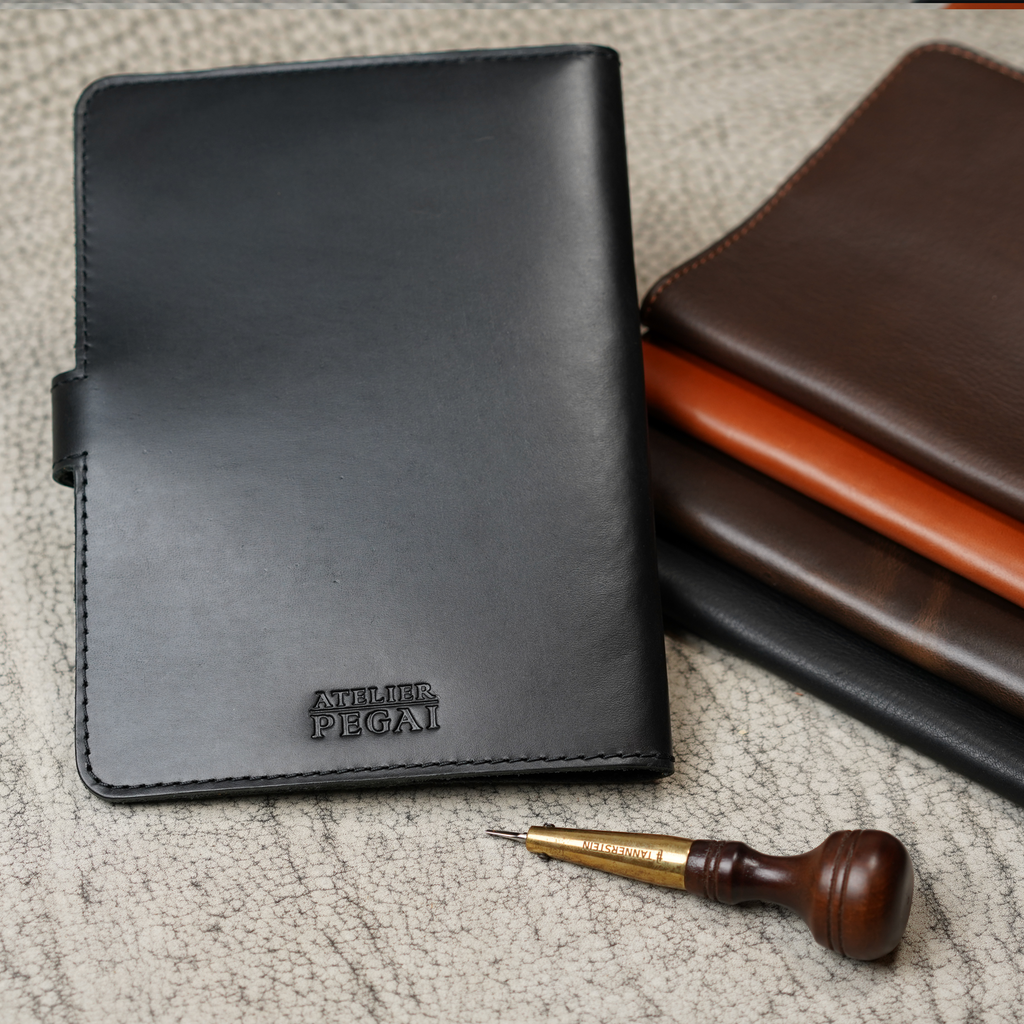 Black DT Lux DuPage journal upright in foreground with embossed Atelier PEGAI stamp, beside a stacked trio of brown-tone DuPage colorways, and a leather awl tool on a textured surface.