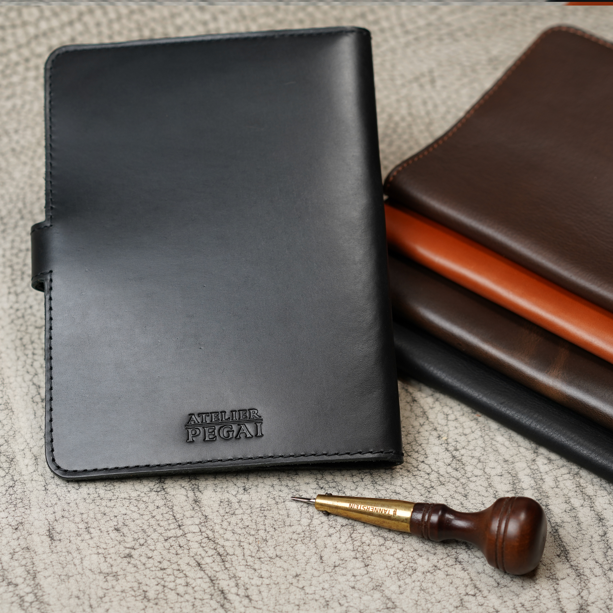 Black DT Lux DuPage journal upright in foreground with embossed Atelier PEGAI stamp, beside a stacked trio of brown-tone DuPage colorways, and a leather awl tool on a textured surface.