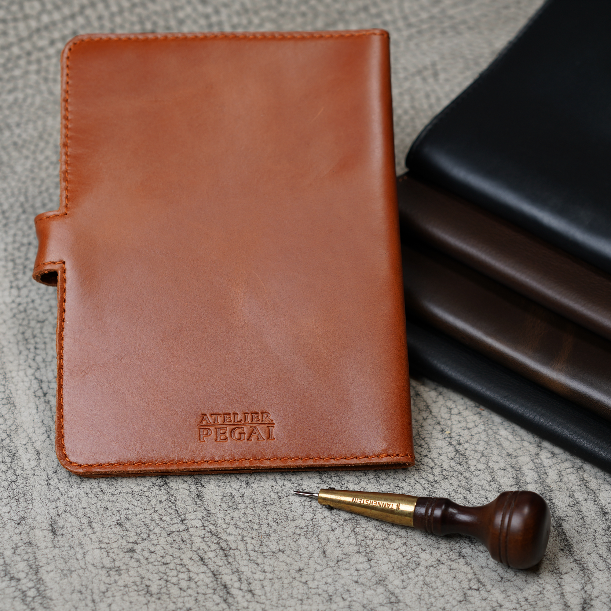 Whiskey Brown DT Lux DuPage journal upright in foreground with embossed Atelier PEGAI stamp, beside a stacked trio of darker DT Lux colorways, and a leather awl tool on a textured surface. 