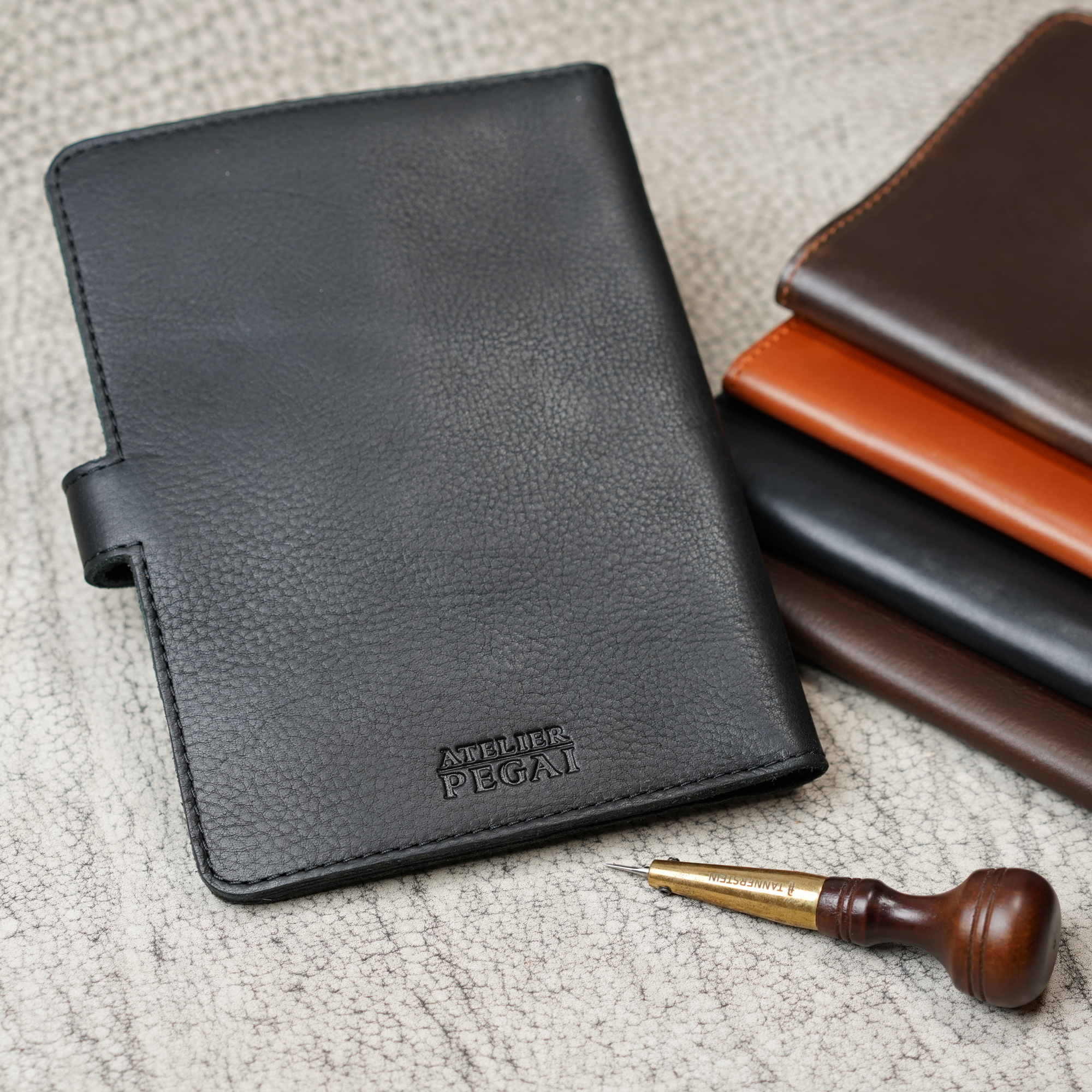 Dymnos Black DuPage journal upright in foreground with embossed Atelier PEGAI stamp, beside a stacked trio of DT Lux colorways, and a leather awl tool on a textured surface.
