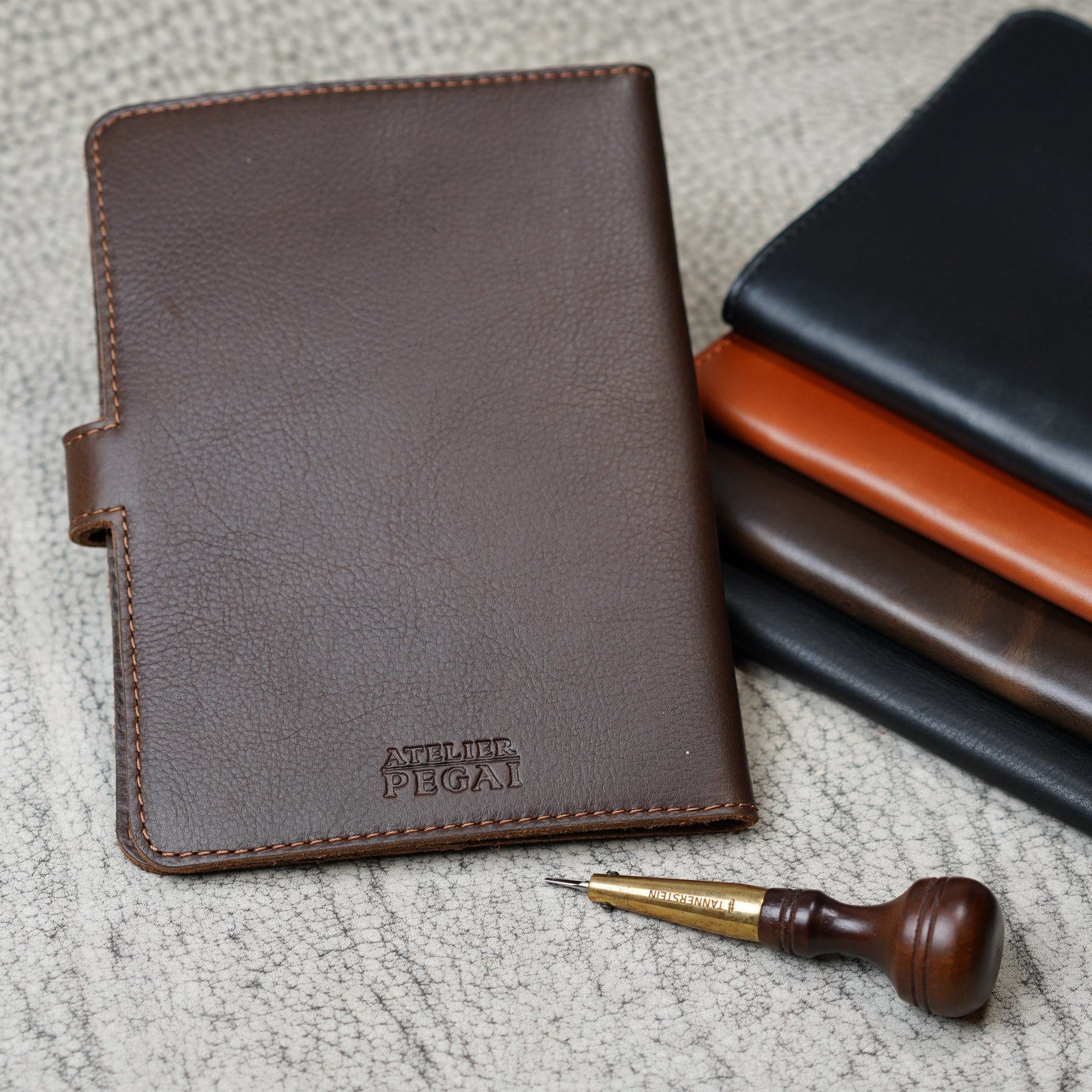 Dymnos Brown DuPage journal upright in foreground with embossed Atelier PEGAI stamp, beside a stacked trio of DT Lux colorways, and a leather awl tool on a textured surface.