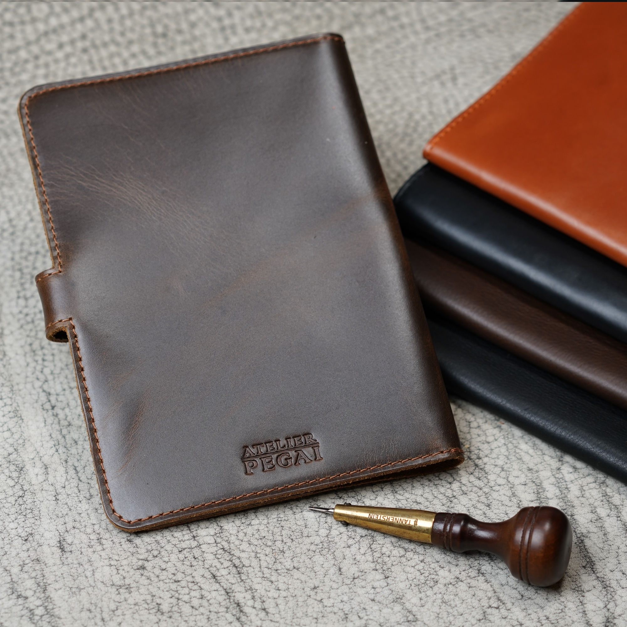Dual Tone Coffee DT Lux DuPage journal upright in foreground with embossed Atelier PEGAI stamp, beside a stacked trio of DT Lux colorways, and a leather awl tool on a textured surface.