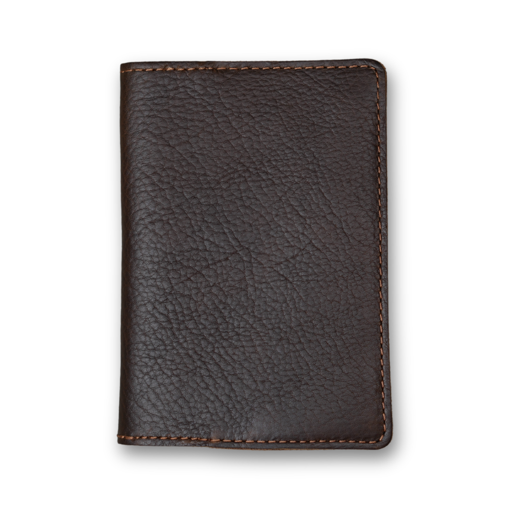Dymnos brown full-grain calfskin passport cover, closed and laid flat. Deep espresso brown pebbled surface with organic grain movement throughout. Hand-stitched edges in contrasting warm brown thread. Rounded corners and clean minimal silhouette with no hardware or closures.
