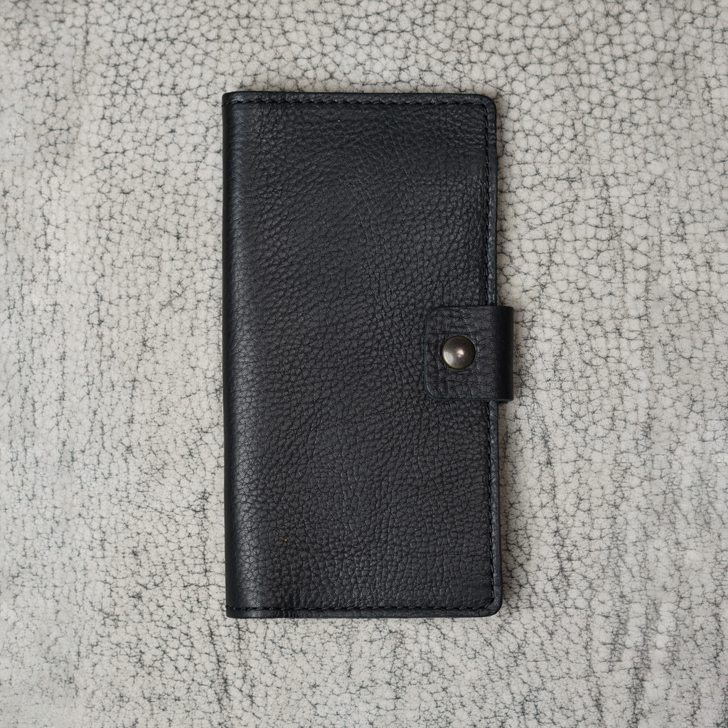 Clark Checkbook Cover in Dymnos Black laid flat on a cream crackled leather surface. Deep black pebbled full-grain Dymnos calfskin with natural grain variation. Antique brass snap on right edge.