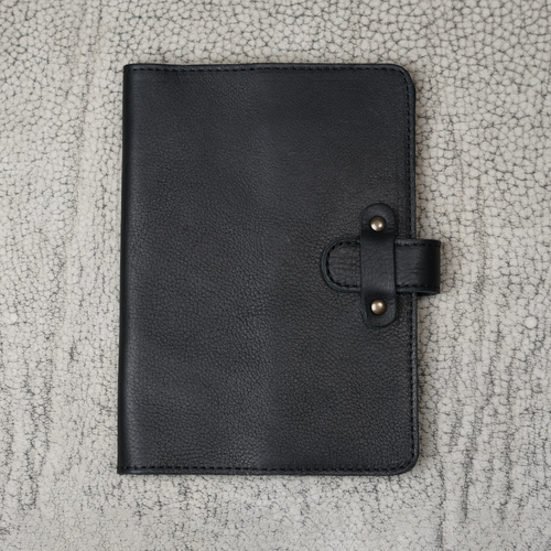 Dymnos Black full-grain calfskin leather journal closed on a cracked stone surface, showing pebbled grain texture, black stitching, and antique brass rivet strap closure.