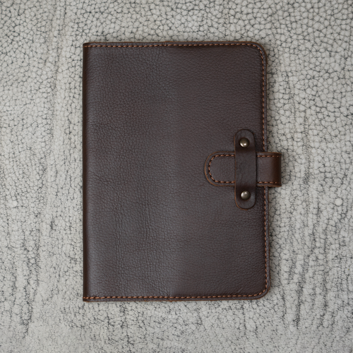 Dymnos Brown top-grain calfskin leather journal closed on a cracked stone surface, showing deep brown pebbled nubuck grain, tan stitching, and antique brass rivet strap closure.