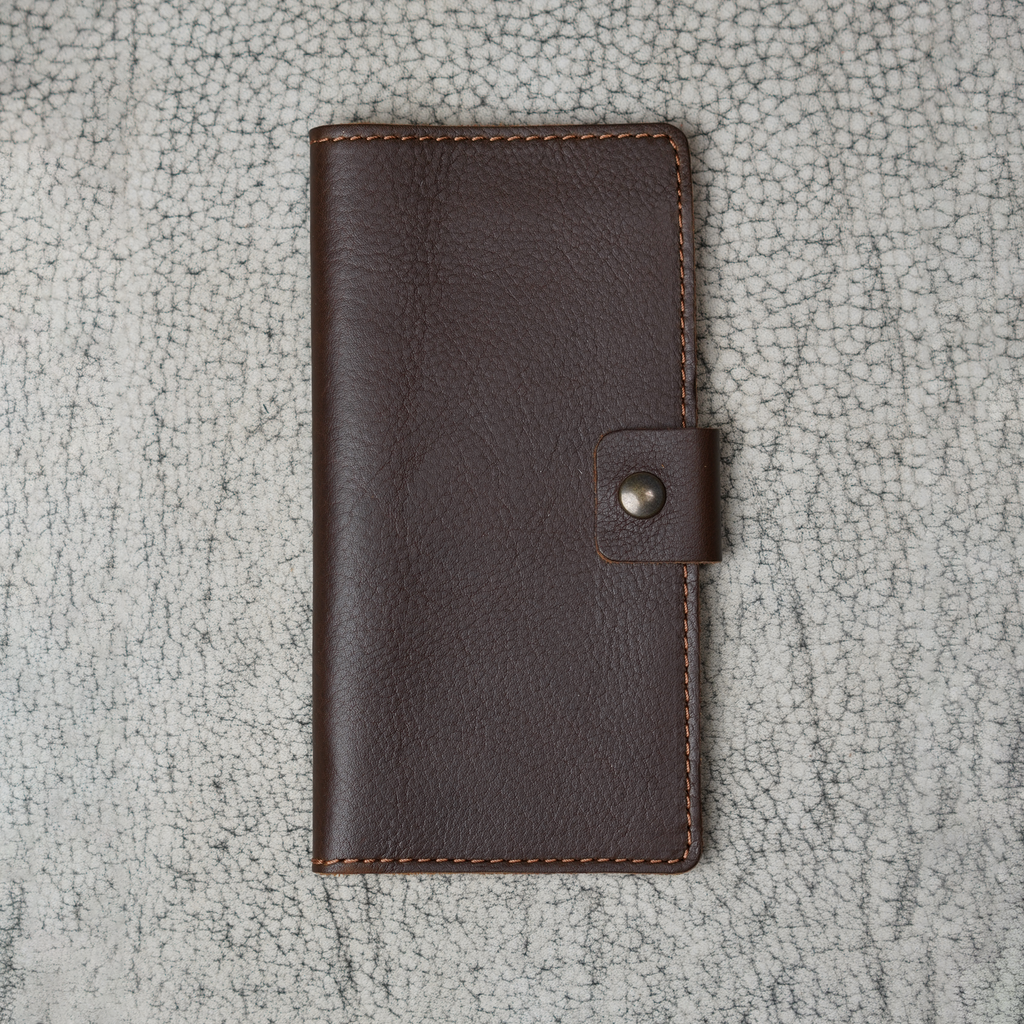 Clark Checkbook Cover in Dymnos Brown laid flat on a cream crackled leather surface. Deep brown pebbled Dymnos full-grain calfskin with natural grain variation. Antique brass snap on right edge.