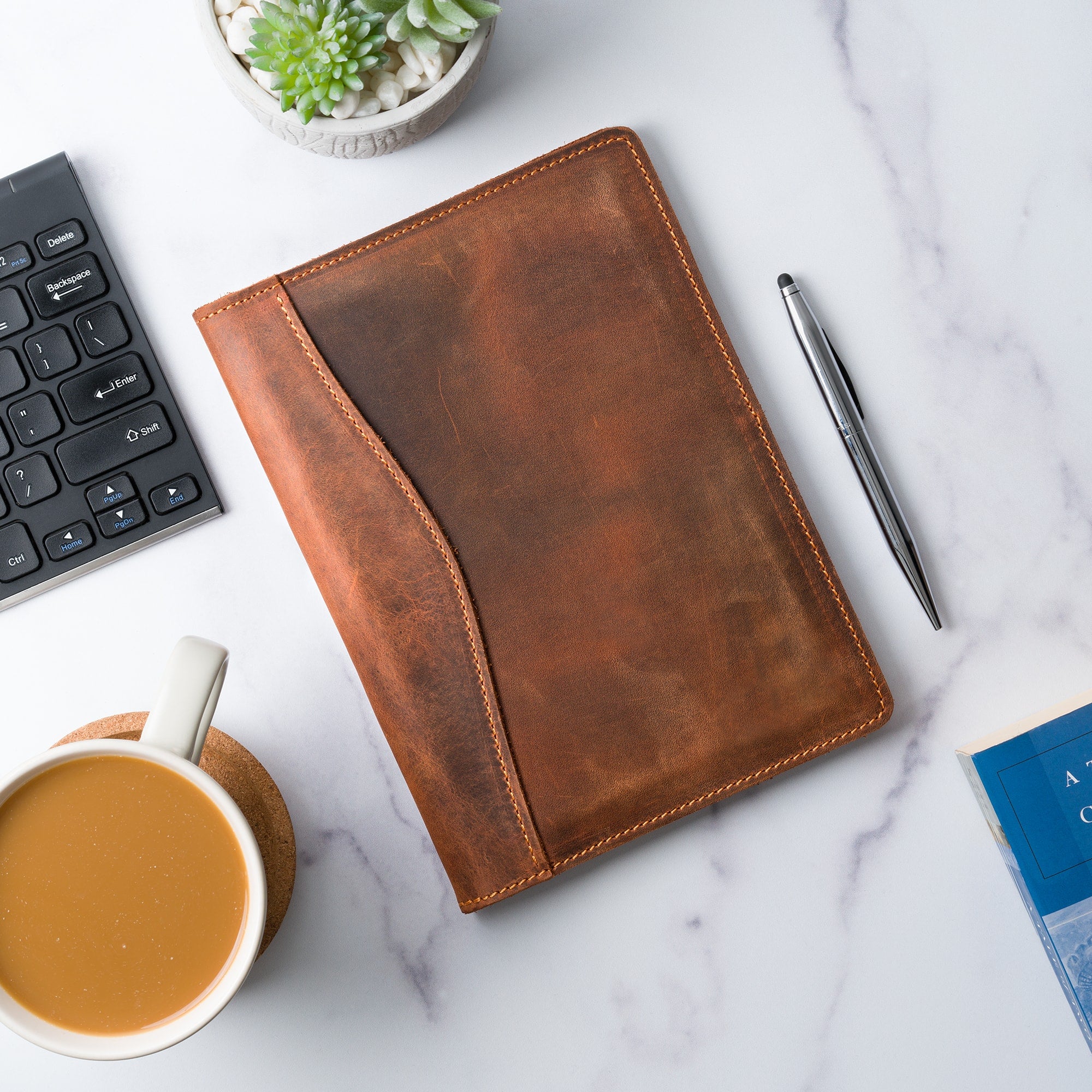 PEGAI Eriksen Jr. padfolio in mahogany Dallas Crazy Horse leather lying closed on a marble desk beside a keyboard, stylus pen, succulent, coffee, and a book.